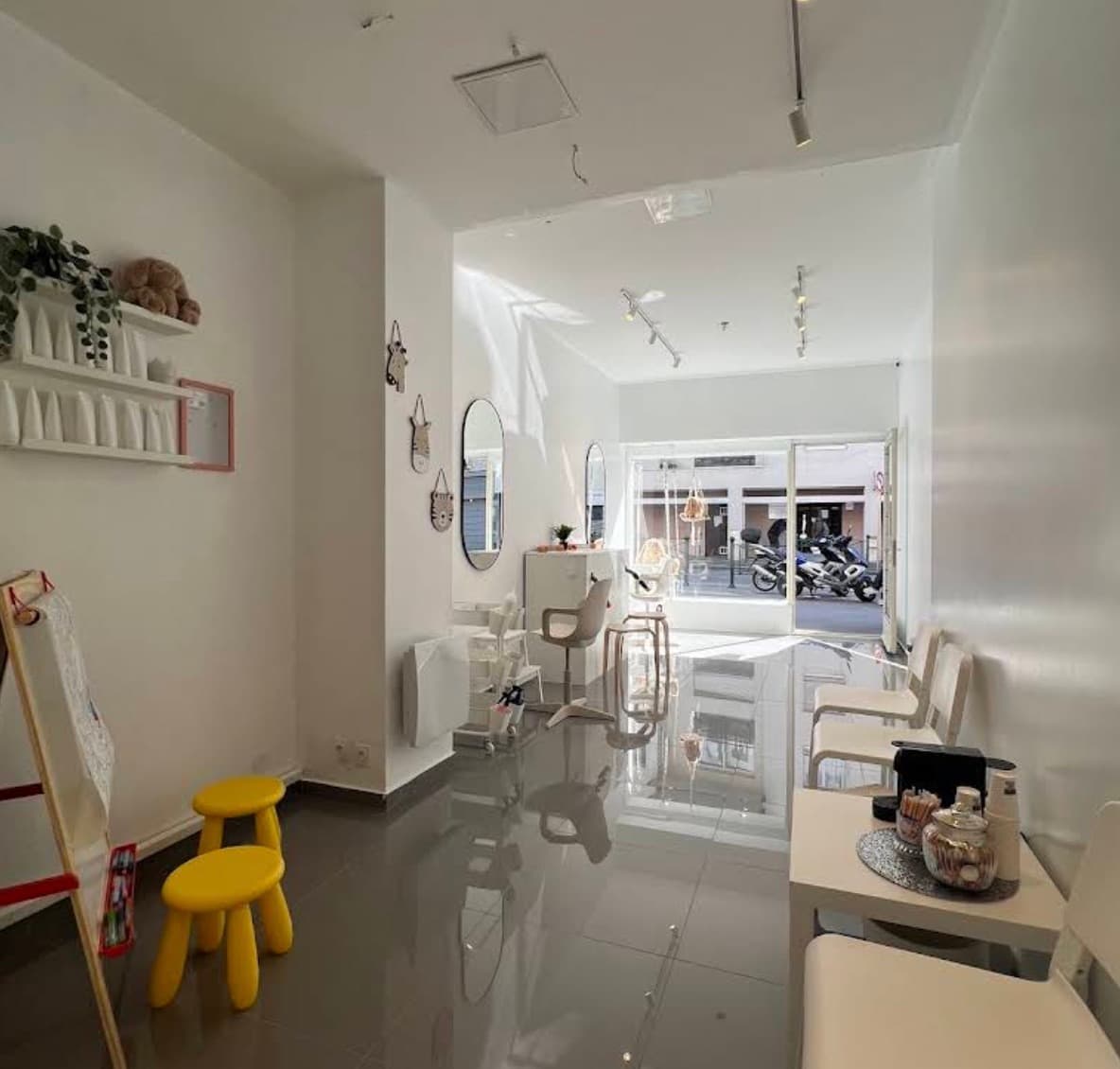 Le coin des enfants : un chevalet à dessin, deux tabourets jaunes, des peluches sur une étagère, et le coin café pour les parents.